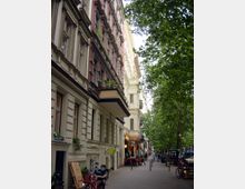 Straßenszene mit mehrstöckigen Altbauten, verziert mit Stuckfassaden und Balkonen, teilweise mit Pflanzen geschmückt. Rechts säumen Bäume die Straße, die von parkenden Fahrrädern, Fußgängern und Café-Tischen belebt ist.