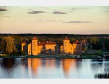 Schloss mit gelber Fassade und roten Dächern, umgeben von einem gepflegten Garten, direkt an einem ruhigen See gelegen, der die Abendsonne reflektiert. Im Hintergrund sind Bäume und eine Häuserreihe zu sehen.
