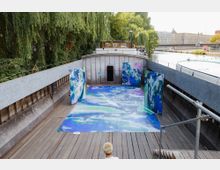 Ausstellung auf einem Holzdeck eines Bootes mit abstrakten Kunstwerken in Blau- und Lilatönen, umgeben von einer Stahlreling. Im Hintergrund sind grüne Bäume, ein Wasserkanal und städtische Gebäude sichtbar.