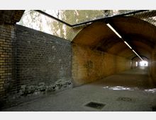 Ein gemauerter Tunnel mit Gewölbedecke, bestehend aus gelben und braunen Ziegeln. Der Boden ist gepflastert, und an der Decke verläuft eine lange Leuchtstofflampe; am Ende des Tunnels ist ein kleines Licht sichtbar.