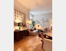 Helles Wohnzimmer mit Holzboden, einer beigefarbenen Eckcouch, einem runden Esstisch und Holzmöbeln. An der weißen Wand befindet sich ein Wandbild eines Baumes, daneben Pflanzen und ein Sideboard mit Dekorationsgegenständen.