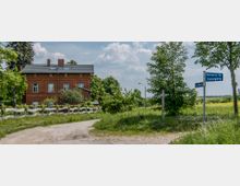 Ein rotes Backsteinhaus in ländlicher Umgebung, umgeben von Bäumen, Wiesen und einem weißen Holzzaun. Im Vordergrund eine unbefestigte Straße mit Wegweisern, die unter anderem "Mehlsdorfer Weg" und "Flämingweg" anzeigen.