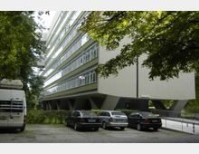 Modernes Büro- oder Wohngebäude mit mehreren Etagen und einer Fassade aus großen Fensterreihen und grünen Streifen. Vor dem Gebäude befinden sich geparkte Autos, umgeben von Bäumen und Grünflächen.