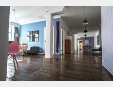 Weitläufiger Innenraum einer Wohnung oder eines Studios mit unbehandeltem Holzfußboden, blauen und weißen Wänden sowie mehreren Sitz- und Dekoelementen. Der Raum ist mit hängenden Lampen, schlichten Möbeln, einem großen Fenster und mehreren Durchgängen ausgestattet.