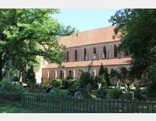 Backsteinkirche im gotischen Stil mit spitzbogigen Fenstern und rotem Ziegeldach, umgeben von einem kleinen, bewachsenen Friedhof mit Grabsteinen und einem Holzzaun. Im Vordergrund stehen Bäume und Büsche, die teils Schatten spenden.