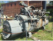 Alte Dampfkesselmaschine aus Metall in einem Freilichtmuseum, umgeben von Kettenabsperrungen, mit einem roten Ziegelgebäude im Hintergrund. Die Maschine zeigt Rostspuren und ist auf einer Wiese ausgestellt.