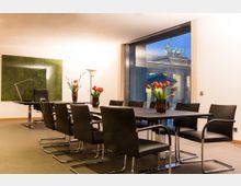Moderner Konferenzraum mit einem großen schwarzen Tisch, umgeben von acht schwarzen Stühlen. Im Raum stehen Vasen mit Tulpen, und durch ein großes Fenster ist das Brandenburger Tor bei Abendbeleuchtung sichtbar.