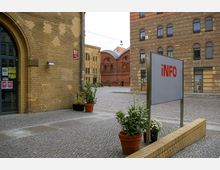 Innenhof mit Klinkergebäuden und gepflastertem Boden, umgeben von restaurierten historischen Fassaden in rot- und beigefarbenen Ziegelsteinen. Im Vordergrund ein "Info"-Schild und mehrere Topfpflanzen.