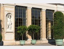 Außenansicht eines historischen Gebäudes mit großen, gitterförmigen Fenstern und einer sandfarbenen Ziegelfassade. Links steht eine Statue in einer Wandnische, davor Pflanzkübel mit Bäumen und Sträuchern.