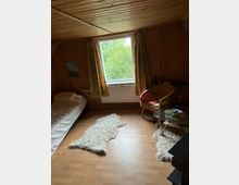 Holzvertäfeltes Zimmer mit einem Einzelbett auf der linken Seite, einem Fenster mit Blick auf Bäume in der Mitte und einem Sessel mit kleinem Glastisch rechts. Der Boden ist aus hellem Laminat, auf dem zwei Schaffell-Teppiche liegen.