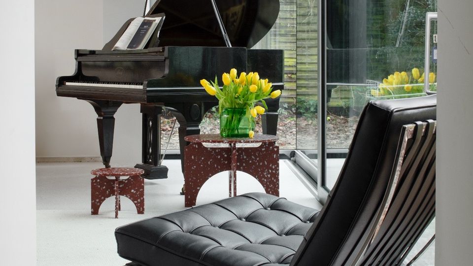 Modern minimalist living room with a black grand piano and a black leather sling chair on a light floor, beside floor-to-ceiling glass walls. A small table with yellow tulips sits near the window, looking out to a garden with trees and a wooden fence.