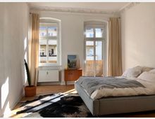 Helles Schlafzimmer mit weißen Wänden, einem Holzboden und zwei großen Fenstern mit beigefarbenen Vorhängen. Das Zimmer enthält ein graues Bett, eine Kommode mit Dekorationen und einen Kaktus in einem Terrakottatopf.