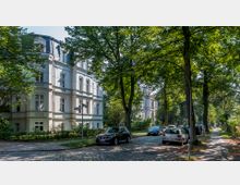Straßenszene in einem Wohngebiet mit historischen, weißen Villen im Gründerzeitstil, umgeben von hohen Bäumen. Kopfsteinpflasterstraße mit parkenden Autos und angrenzendem Gehweg im Schatten.
