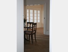 Innenraumansicht mit einem Holzboden in Fischgrätmuster, einem eleganten Holztisch mit Stühlen und einer weißen Doppeltür mit Glaseinsätzen im Hintergrund. Der Raum wirkt hell durch die helle Wandfarbe und natürliche Beleuchtung.