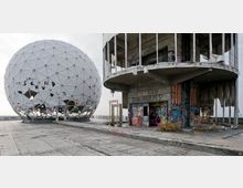 Eine verlassene Radaranlage mit einer großen, weiß verkleideten Kuppelstruktur und einem angrenzenden, teils zerstörten Betongebäude, das mit Graffiti bedeckt ist. Der Boden ist aus Betonplatten und weist ebenfalls Spuren von Verwitterung und Graffiti auf.