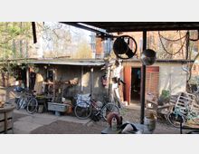 Hinterhof mit improvisierter Werkstatt und Lagerbereich, umgeben von einfachen Schuppen und Fahrrädern. Im Vordergrund sind Werkzeuge, Fahrräder und Alltagsgegenstände verteilt, während im Hintergrund ein Gebäude aus Ziegelstein sichtbar ist.