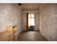 Schmaler Raum mit unverputzten Wänden und einer Decke mit sichtbaren Gebrauchsspuren. Ein einfaches Holzregal sowie eine Kommode stehen links, während das Ende des Raums von einem Fenster mit gelbem Vorhang und einem Heizkörper geprägt ist.