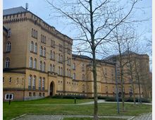 Historisches Backsteingebäude mit mehreren Stockwerken, zahlreichen Rundbogenfenstern und verzierten Fassaden, umgeben von gepflasterten Wegen, kahlen Bäumen und einer Rasenfläche. Der Himmel ist bewölkt.