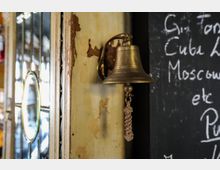 Messingfarbene Glocke an einer Wand mit abblätternder Farbe, daneben eine Tafel mit handgeschriebener Schrift. Links befindet sich ein Bereich mit verzierten Glaselementen.