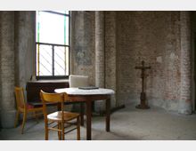 Ein einfacher Raum mit rohen Backsteinwänden und einer großen, farbig verglasten Fensterscheibe. Im Vordergrund stehen ein runder Holztisch mit einer weißen Tischdecke, zwei Stühle und eine braune Kommode, während an der Wand ein Holzkreuz auf einem Sockel steht.