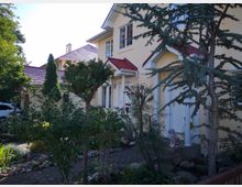 Ein zweistöckiges gelbes Wohnhaus mit roten Dächern, flankiert von einem gepflegten Garten mit Pflasterweg, verschiedenen Bäumen, Sträuchern und dekorativen Steinen. Vor dem Eingang steht eine kleine Löwenstatue.