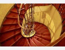 Geschwungene Wendeltreppe mit rotem Teppichbelag, dekorativem Geländer aus Schmiedeeisen und einer Wand mit gelbem, floralen Muster. Die Treppe erzeugt durch das Licht Schattenspiele auf der Wand.