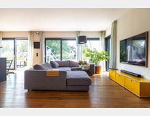 Wohnzimmer mit großer grauer Eckcouch, gelbem Lowboard und Flachbildfernseher an der Wand. Große Fensterfront mit transparenten Vorhängen bietet Blick auf einen Garten, durchflutet den Raum mit Tageslicht und ist von Holzboden ergänzt.