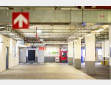 Ein unterirdisches Parkhaus mit hellen Wänden, mehreren weißen Säulen und einer roten Beschilderung für die "Ausfahrt". Im Hintergrund befinden sich ein roter Ticketautomat, Türen und ein grünes Notausgangszeichen. Die Decke ist mit sichtbaren Lüftungsschächten und Kabeln ausgestattet.