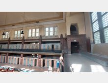 Innenansicht eines historischen Schwimmbades mit mehreren Umkleideräumen an den Wänden, großen Fenstern mit Glasbausteinen und Ziegelmauerwerk. Sichtbare Deckenbeleuchtung und bauliche Details wie Geländer und ein offener Durchgang ergänzen die Szenerie.