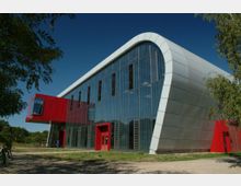 Modernes Gebäude mit geschwungener, metallverkleideter Fassade und großen Glasflächen. Ein auffälliger, roter Anbau ragt seitlich aus dem Baukörper heraus, umgeben von Bäumen und einer Grasfläche.