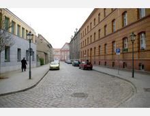 Kopfsteinpflasterstraße mit geparkten Autos, flankiert von einem historischen Gebäude aus gelbem und rotem Backstein auf der rechten Seite und modernen Gebäuden sowie älteren Bauwerken auf der linken Seite; im Hintergrund ein rosa Gebäude mit Giebel. Straßenlaternen und Gehwege sind sichtbar.