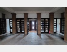 Eingangsbereich eines Gebäudes mit Säulen aus beigem Stein und dunklen Holztüren mit Glaseinsätzen. Im Hintergrund ist eine mittig platzierte Holztür mit horizontalen Fensterausschnitten, die von natürlichem Licht durchflutet wird.