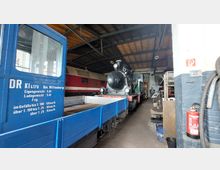 Ein Innenraum eines Bahndepots mit einer alten Dampflok und einer roten Diesellok nebeneinander. Im Vordergrund steht ein blauer Transportwagen mit aufgedruckten technischen Daten, während an der rechten Wand ein Feuerlöscher hängt.