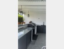 Überdachte Außenküche auf einer Terrasse mit weißen Deckenbalken und hängenden Laternenleuchten, ausgestattet mit dunklen Unterschränken und grauer Arbeitsplatte. Im Vordergrund stehen ein Gasgrill und rechts eine Spüle; im Hintergrund sind Pflanzen und Sitzmöbel zu sehen.