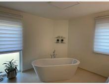 Modernes Badezimmer mit freistehender Badewanne in der Mitte, zwei Fenstern mit halbtransparenten Rollos und einem Regal mit Dekorationen an der Wand. Links unten steht eine Zimmerpflanze in einem silbernen Topf auf dem Boden.