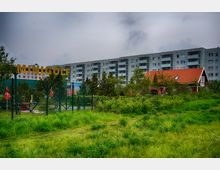 Grünfläche mit einer umzäunten Minigolf-Anlage im Vordergrund, darüber ein gelbes Schild mit der Aufschrift „MINIGOLF“. Im Hintergrund befinden sich ein Wohnblock aus grauen Mehrfamilienhäusern sowie ein rot gedecktes Gebäude neben Bäumen.