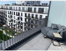 Terrasse mit Geländer und moderner Sitzgruppe, bestehend aus einer grauen Rundcouch und einem kleinen Beistelltisch. Im Hintergrund sind ein mehrstöckiges Wohngebäude mit Balkonen sowie ein Innenhof mit Grünflächen zu sehen.