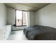 Ein modernes Schlafzimmer mit minimalistischer Einrichtung. Das Zimmer hat eine große Fensterfront mit weißen Vorhängen, einen weißen Schrank, einen schwarzen Stuhl und ein Bett mit schwarzer, gemusterter Bettdecke.