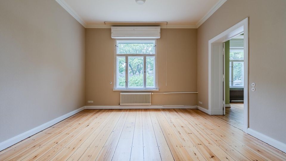 Leerer Wohnraum mit hellem Holzdielenboden, beigefarbenen Wänden und weißer Stuckleiste an der Decke. Mittig ein großes Fenster mit Heizkörper darunter und rechts eine offene Tür zu einem angrenzenden Zimmer.