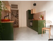 Schmale Küche mit grünen Unterschränken, einer Arbeitsplatte aus Holz, und einer Rückwand mit rosa Fliesen. Im Hintergrund ist ein Abstellraum mit zwei aufrechten Fahrrädern zu sehen, und rechts im Bild steht ein Holzbogenstuhl an einem kleinen runden Tisch.