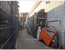 Enger, gepflasterter Weg zwischen einer hohen Metallgitterzaunwand links und einer weißen Backsteinmauer rechts. Rechts stehen Regale mit Abdeckplanen und Autoteilen, darunter eine leuchtend orangefarbene Autotür. Im Hintergrund sind Bäume und ein Fahrzeugteil sichtbar.