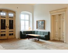 Heller Innenraum mit hohen weißen Wänden, großen Flügeltüren und einem hohen Fenster in einer Nische. In der Ecke steht ein L-förmiges grünes Sofa mit niedrigem Couchtisch; an der Wand hängt ein gerahmtes Bild, der Boden ist teils mit alten Fliesen ausgelegt.