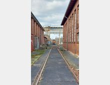 Schmalspur-Gleisanlage zwischen zwei langen Ziegelgebäuden mit großen Fenstern, im Hintergrund eine orange Lokomotive und eine Überführung aus Metall. Die Umgebung wirkt industriell mit teils bewachsenen Schienen und grauem Himmel.