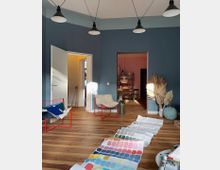 Wohnraum mit dunkelblau gestrichenen Wänden, Holzboden und mehreren modernen Einrichtungsgegenständen. Im Vordergrund liegen auf dem Boden große Blätter mit Farbmusterkarten, im Hintergrund sind eine offene Tür zu einem weiteren Raum und dekorative Elemente wie getrocknete Palmblätter sichtbar.