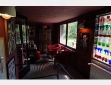 Kleiner Raum mit roten Wänden und einer gemütlichen Einrichtung, darunter zwei rote Sessel, ein kleiner runder Tisch und eine bunt gemusterte Teppichmatte. Große Fenster bieten Blick auf einen Garten, während ein Regal mit farbigen Gläsern und Wanddekorationen den Raum schmücken.