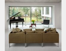 Minimalist living room with a grand piano, floor lamp, and low tables in front of a large wall of sliding glass windows overlooking a garden. A sectional sofa faces the windows, with a black leather lounge chair near the glass.