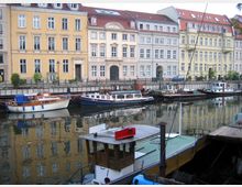Mehrere kleine Boote und Yachten liegen in einem ruhigen Kanal vor einer Reihe historischer, pastellfarbener Wohnhäuser mit roten Ziegeldächern. Im Vordergrund ist der Bug eines größeren Boots zu sehen, das teilweise ins Bild ragt.