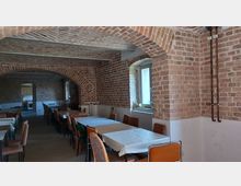 Ein Raum mit unverputzten Ziegelwänden und gemauerten Bögen, eingerichtet mit mehreren Tischen und Stühlen. Im Hintergrund führt eine Türöffnung zu einer kleinen Küche, und ein Fenster lässt natürliches Licht herein.