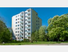 Mehrstöckiges Wohngebäude mit Balkonen, umgeben von Bäumen und Grünflächen, bei klarem, sonnigem Himmel. Im Vordergrund befindet sich eine Wiese und ein Fußweg.