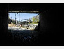 Blick aus einem dunklen Tunnel auf einen Bahnsteig mit Überdachung, Schienen und Signalstruktur. Im Vordergrund ist eine Kopfsteinpflasterstraße, rechts ein Bauzaun, und links ein Sandhaufen sichtbar.